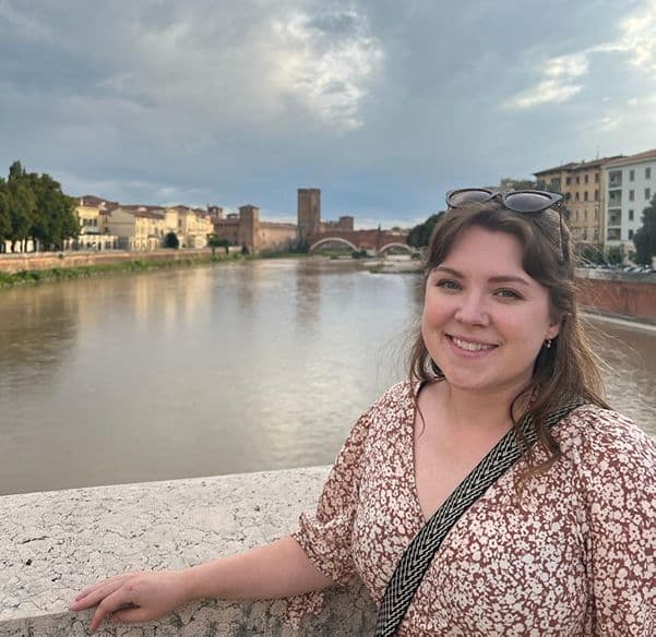 Lady wearing brown and white dress stood in front of river and buildings in the background